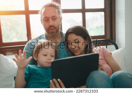 Happy family, Little baby boy with mom and dad lying on bed together. while looking tablet computer online internet watching cartoon, social media enjoy together during holiday at home.