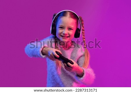 Happy sweet blondy preteen girl playing video games on colorful background in neon light, using wireless headphones and holding joystick. Game addiction for children concept