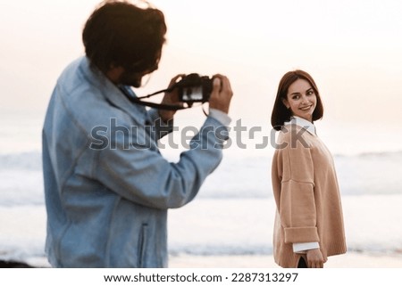Millennial middle eastern husband taking photo on camera of caucasian female on ocean beach. Weekend walk, relax and rest together, vacation, date at nature, blog and social network