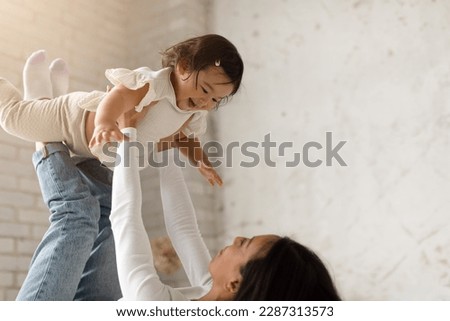 Cheerful Chinese Mommy Playing With Adorable Infant Daughter, Lifting Child Riding Her On Legs Lying In Modern Bedroom At Home. Mother Having Fun Cuddling With Her Little Toddler Baby