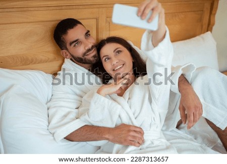 Happy Couple Using Smartphone Making Selfie Posing Wearing White Bathrobes Lying In Bedroom In Hotel Indoors. Spouses Enjoying Honeymoon Taking Photos Having Fun On Vacation. Selective Focus