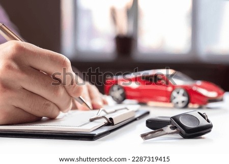 Car keys on desk with man signing purchase documents in background. Closeup of black modern car keys while hand complete the insurance policy or rental documents. Guy buying new car at dealership.