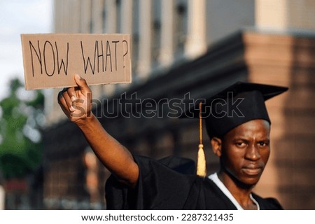 Portrait closeup of black guy holding in hand upraised cardboard poster on street looking for job, soft focus. Hiring and employment issue. University or college graduating student in gown and cap. Royalty-Free Stock Photo #2287321435