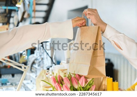 unrecognizable female vendor hands over a paper bag filled with purchases to a customer at a vibrant marketplace, showcasing the growing popularity of environmentally conscious shopping. Royalty-Free Stock Photo #2287345617