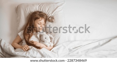 Cute funny cheerful little girl with brown hair sleeping in the white bed with favorite fluffy toy bunny and near lying clock. Horizontal banner. Copy space. . High quality photo