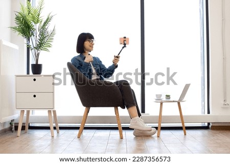 Young brunette female in casual clothes presenting thumb-up sign while posing at cell phone camera on selfie stick in office interior. Relaxed worker taking photo in soft armchair near coffee table.