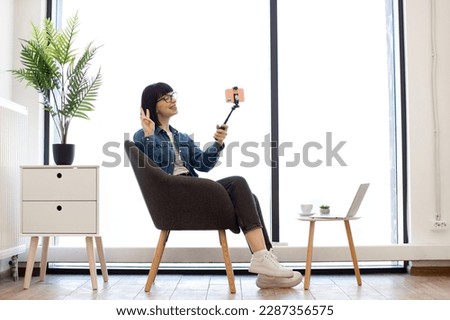 Beautiful young woman in denim jacket holding selfie stick with smartphone while relaxing in office during coffee break. Smiling female worker taking self-portrait with victory sign in cozy workplace. Royalty-Free Stock Photo #2287356575