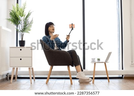 Beautiful young woman in denim jacket holding selfie stick with smartphone while relaxing in office during coffee break. Smiling female worker taking self-portrait with victory sign in cozy workplace.