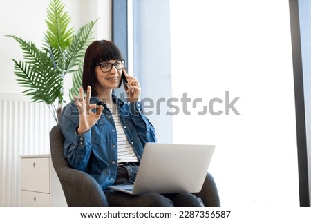 Happy caucasian woman in casual clothes talking on smartphone while surfing internet via laptop during coffee break in office showing sign ok.