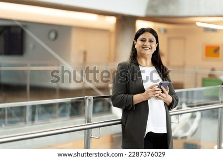 Smiling attractive confident Indian woman holding mobile phone looking at camera, copy space. Technology concept Royalty-Free Stock Photo #2287376029