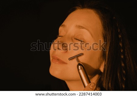 Young beautiful white woman, girl is using vibrating golden facial massager on dark black background with copy space. Preservation prolongation of natural beauty youth Female skin care with pleasure.  Royalty-Free Stock Photo #2287414003