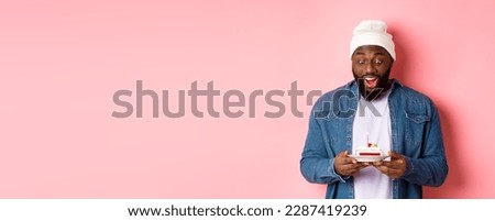 Cheerful african-american guy celebrating birthday, making wish on bday cake with lit candle, smiling happy, standing over pink background.