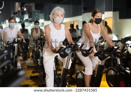 Portrait of young and senior women wearing face mask for disease protection training together during stationary bike workout in gym