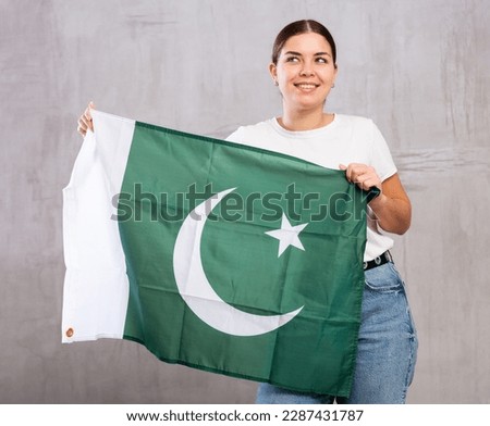 Happy young woman in casual clothes holding flag of Pakistan against gray wall indoors