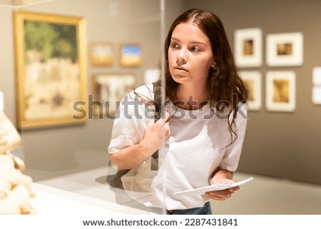 Attentive girl visitor looks with interest at the exhibit in the museum, located behind the glass Royalty-Free Stock Photo #2287431841