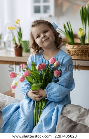 Cute little girl, baby daughter in a blue dress prepare pink tulip flowers to her happy mom. Celebration of Mother's or Women's day, birthday or anniversary. Cozy home atmosphere, kitchen. Springtime