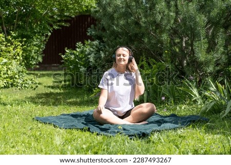 A young blonde girl is sitting on the grass and listening to music in wireless headphones.