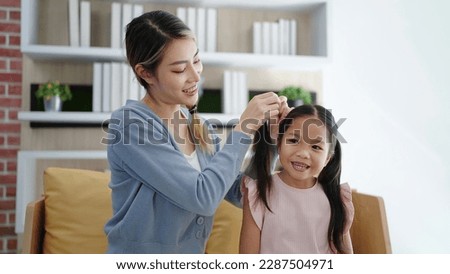 Happy loving family Asian mother is combing her daughter's braiding hair sitting on sofa at living room. Anti lice treatment