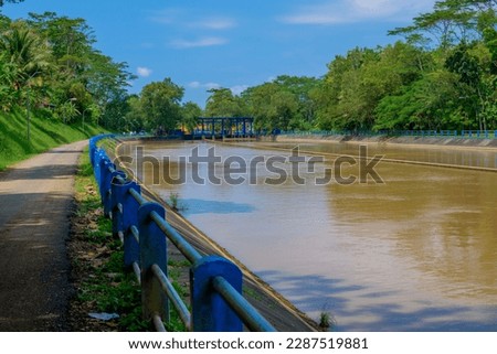 Scenery of the asphalt road by the river with green trees all around. Among them there is a blue guardrail to make it safer. This river in Kebasen, Banyumas is brown in color.