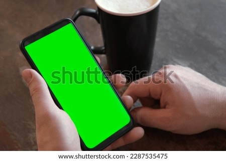 Man Use of Mobile Phone with Blank Green Screen in a Cafe on Rustic Vintage Wooden Table. Male Holding Smart Phone Empty ChromaKey Screen Mock up in Coffee Shop. Morning. Showing Mock up.