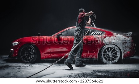 Car Wash Specialist Using Pressure Washer to Rinse a Red Modern Sportscar. Adult Man Washing Away Dirt, Preparing a Tuned Car for Detailing. Creative Cinematic Photo in Studio Royalty-Free Stock Photo #2287564621