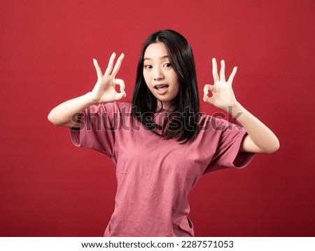 A studio portrait of a young Indonesian (Asian) woman wearing a pink shirt and displaying the OK sign with her hand. Isolated with a red background.