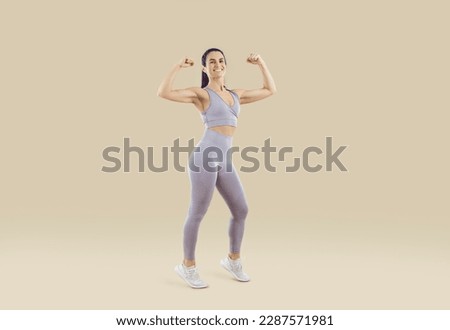 Beautiful sporty woman showing off athletic body and muscular arms on beige background. Full length portrait of fitness woman in sportswear showing biceps on camera. Workout sport motivation concept. Royalty-Free Stock Photo #2287571981