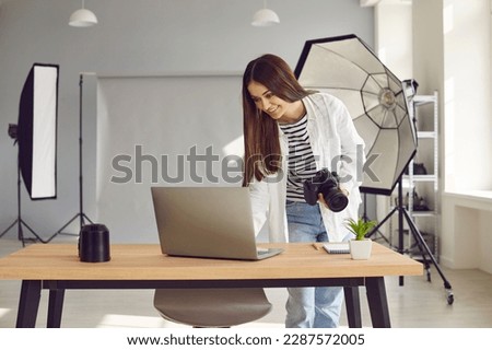 Portrait of a young happy smiling woman photographer with camera and lenses on her workplace looking cheerful at the laptop monitop screen in production studio with professional equipment.