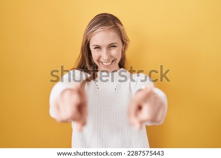 Young caucasian woman wearing white sweater over yellow background pointing to you and the camera with fingers, smiling positive and cheerful 