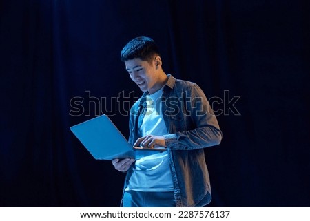 Young guy, student looking on laptop, working online, posing against dark background in neon light. Modern technologies. Concept of human emotions, youth, fashion, lifestyle, education