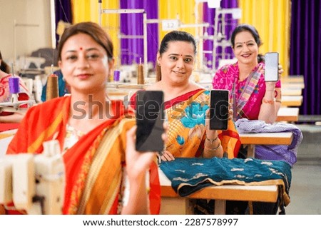 Indian women showing smartphone screen while working on sewing machine at factory