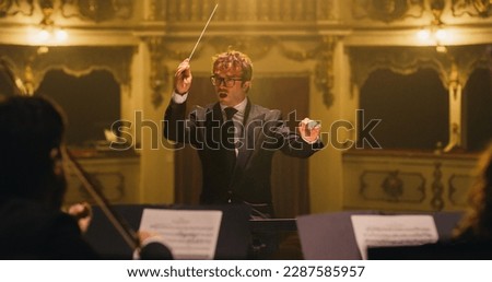 Cinematic Close Up of Conductor Directing Symphony Orchestra with Performers Playing on Stage During Music Concert. Professional Conductor Leading Musicians Passionately in Classic Theater Royalty-Free Stock Photo #2287585957