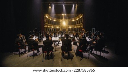 Wide Cinematic shot of Conductor Directing Symphony Orchestra with Performers Playing Violins, Cello and Trumpet on Classic Theatre with Curtain Stage During Music Concert. Back View Royalty-Free Stock Photo #2287585965