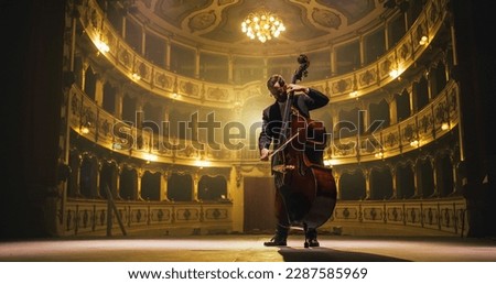 Cinematic shot of Male Cellist Playing Cello Solo on an Empty Classic Theatre Stage with Dramatic Lighting. Professional Musician Rehearsing Before the Start of a Big Show with Orchestra Royalty-Free Stock Photo #2287585969