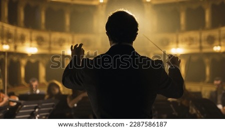 Back View Cinematic shot of Conductor Directing Symphony Orchestra with Performers Playing Violins, Cello and Trumpet on Classic Theatre with Curtain Stage During Music Concert Royalty-Free Stock Photo #2287586187