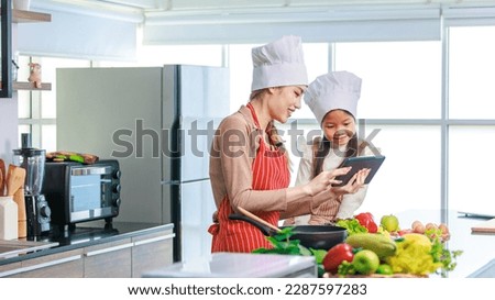 Asian young female chef housewife mother wears white tall cook hat and apron showing mockup glass touchscreen tablet computer screen to little cute girl chef learning cooking online at home kitchen.