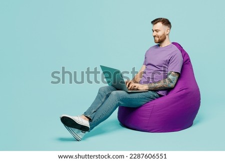 Full body smiling happy young IT man he wears purple t-shirt sit in bag chair hold use work on laptop pc computer isolated on plain pastel light blue cyan background studio portrait. Lifestyle concept