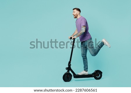 Full body side view caucasian smiling fun happy young man he wearing purple t-shirt riding e-scooter raise up leg isolated on plain pastel light blue cyan background studio portrait. Lifestyle concept Royalty-Free Stock Photo #2287606561