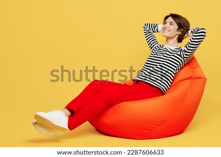 Full body minded fun calm happy young woman wear casual black and white shirt sit in bag chair hold hands behind neck dream isolated on plain yellow color background studio portrait. Lifestyle concept