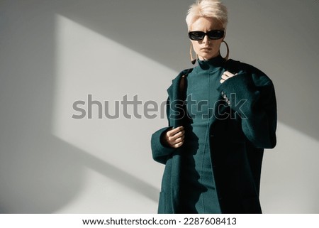 young woman in trendy sunglasses standing in green dress and coat on grey background
