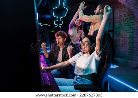 Group of excited friends celebrating victory of young asian girl in online video game while standing next to her in modern cybersport club