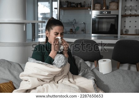 Ill upset young woman sitting on sofa covered with blanket freezing blowing running nose got fever caught cold sneezing in tissue, sick girl having influenza symptoms coughing at home, flu concept Royalty-Free Stock Photo #2287650653