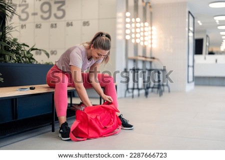 Beautiful satisfied athletic young woman in sportswear sitting on bench in locker room at gym, packing sports equipment in gym bag after finished workout fitness training session, preparing to go home