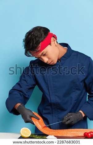 Focused young man in blue chef's jacket working inside the studio, going to cook delicious fish dish with fillet of salmon, tasty food concept, copy space, high quality photo