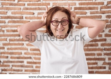 Senior woman with glasses standing over bricks wall posing funny and crazy with fingers on head as bunny ears, smiling cheerful 