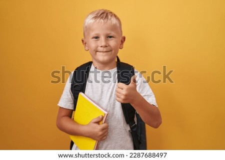 Little caucasian boy wearing student backpack and holding book smiling happy and positive, thumb up doing excellent and approval sign 