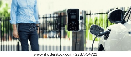 Focus closeup electric vehicle recharge battery at public charging station in the city area with blur businessman in background. EV car attached with electric charger for eco-friendly idea.