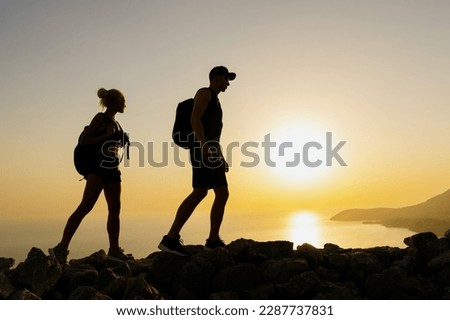A man and a woman walk on rocks in the mountains against the sunset. Silhouettes of tourists