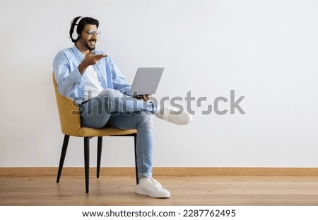 Happy young middle eastern guy in casual and glasses, wireless headphones has video call on laptop on gray wall background. Watch video for work, business, communication remotely at home