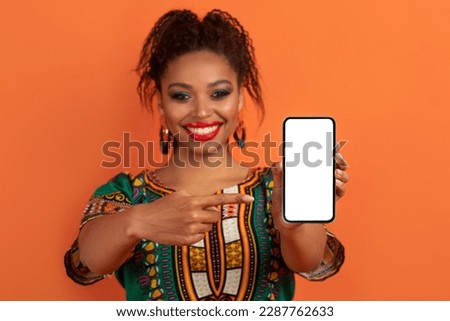 Positive cheerful smiling pretty young black woman wearing bright makeup and traditional african costume showing modern cell phone with white blank screen, mockup for online offer, orange background